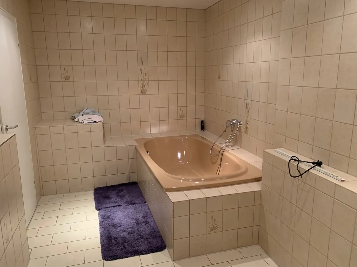 Altes Badezimmer mit beigen Fliesen, Eckbadewanne und lila Badvorleger